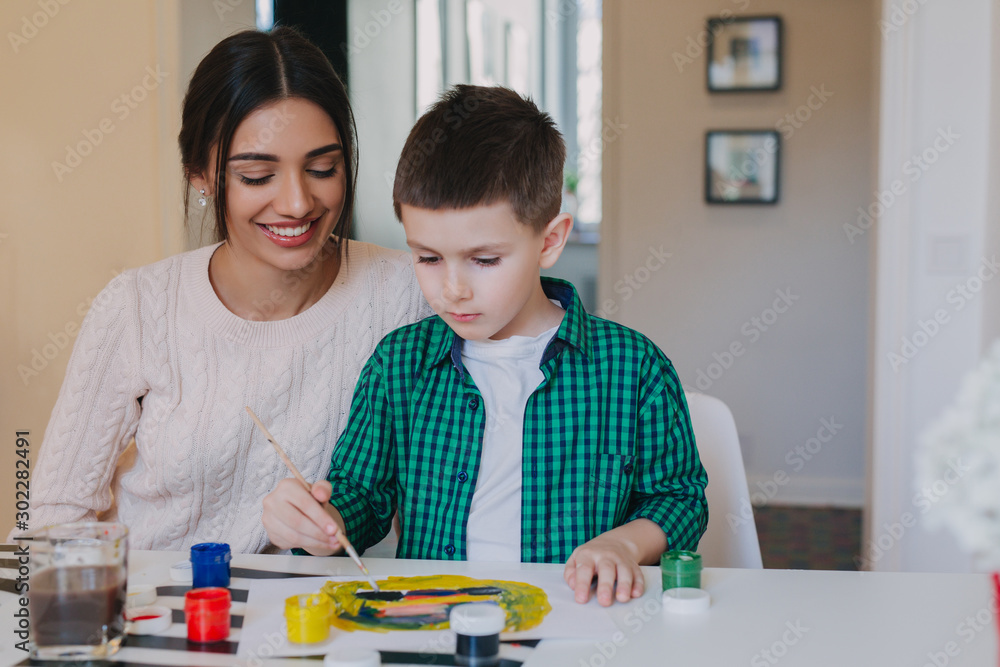 Mother and little son together paint at home. Woman helps her child