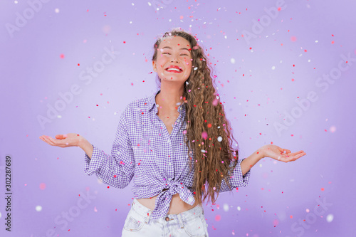 Portrait of a beautiful girl in a rain of confetti shot over an isolated purple background. Girl with closed eyes smiles widely.