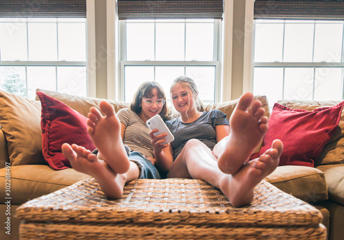 Two teenage girls sitting on couch with feet up looking at cellphone. photo