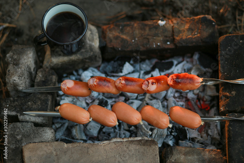Sausages on a skewer in the forest. Camping with grilled sausages and coffee. Charcoal grill picnic