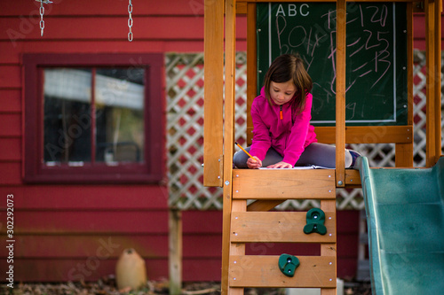 A little girl sits in a playset writing in a notebook with a pencil