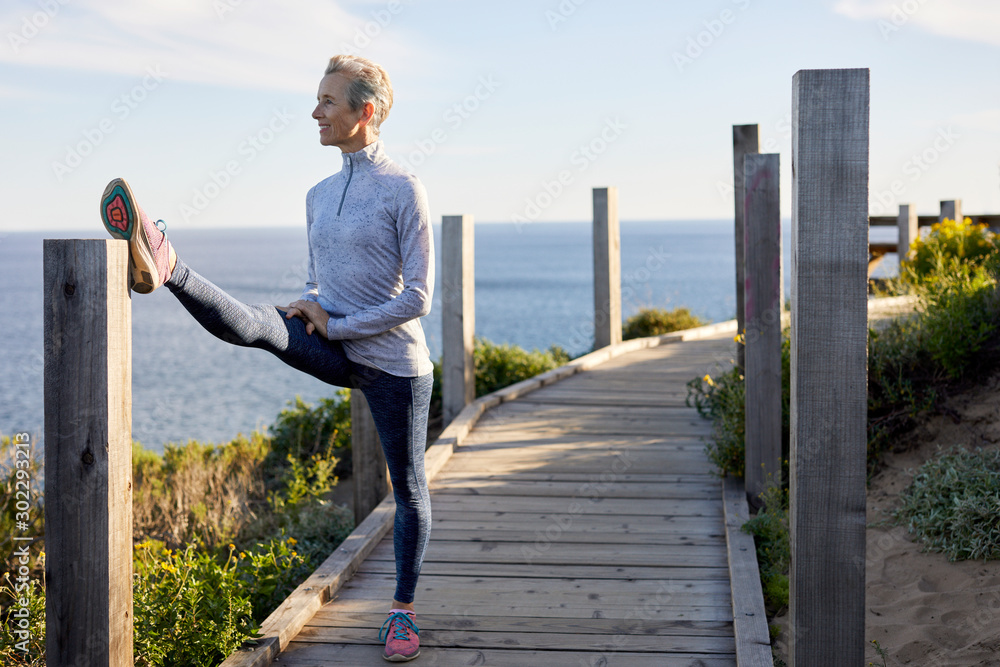 Full length of senior woman doing stretching while standing on boardwalk by sea against sky Full length of senior woman doing stretching while standing on boardwalk by sea against sky