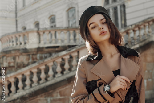 Outdoor autumn fashion portrait of young elegant lady wearing trendy faux leather beret, classic beige trench coat, wrist watch, posing in street of European city. Copy, empty space for text