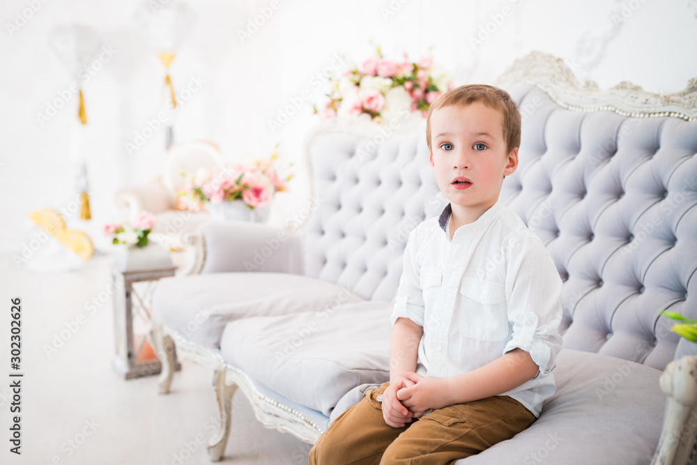 Funny nosy little boy sits on a blue sofa in a beautiful cozy room and looks somewhere. The concept of naive little children. Child care concept Funny nosy little boy sits on a blue sofa in a beautiful cozy room and looks somewhere. The concept of naive little children. Child care concept