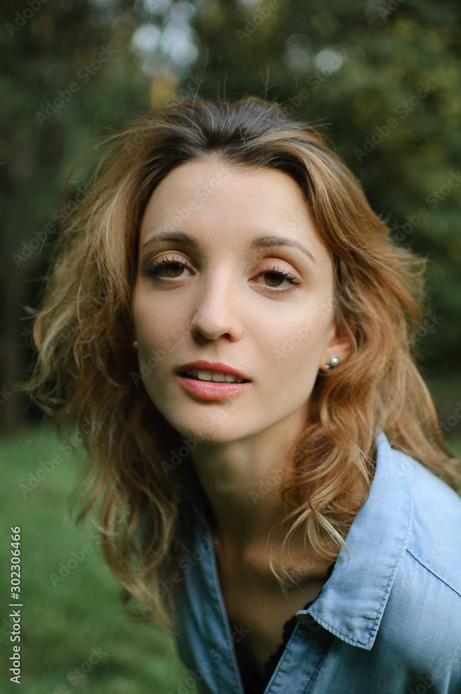 Blonde girl wearing blue casual jeans blouse is looking at the camera in the park on green trees background Blonde girl wearing blue casual jeans blouse is looking at the camera in the park on green trees background