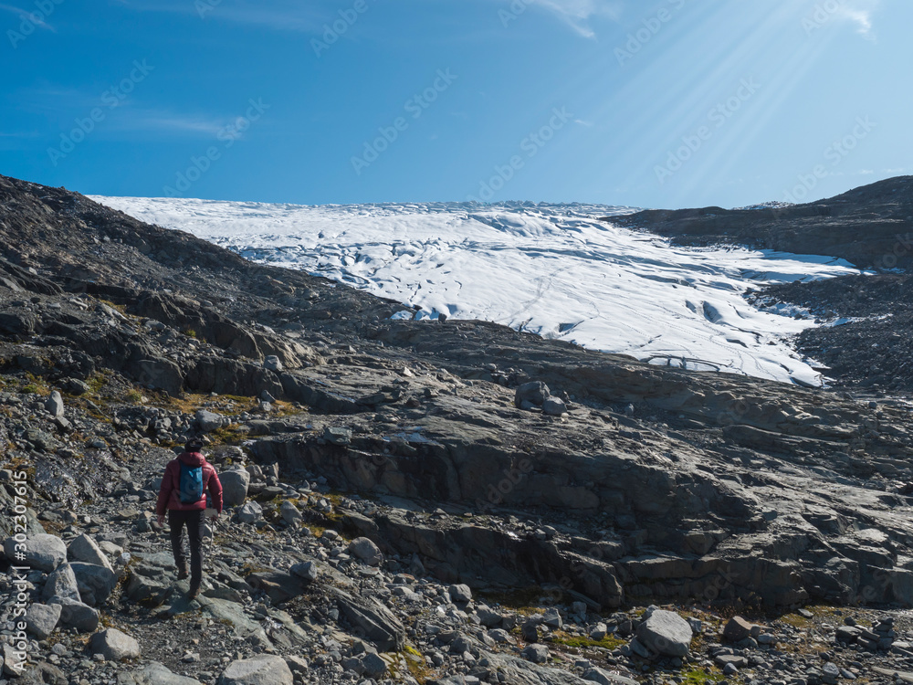 Man hiker in red down jacket on hiking trail to glacier Smorstabbreen in Jotunheimen National Park, Norway. Sunny day, blue sky background