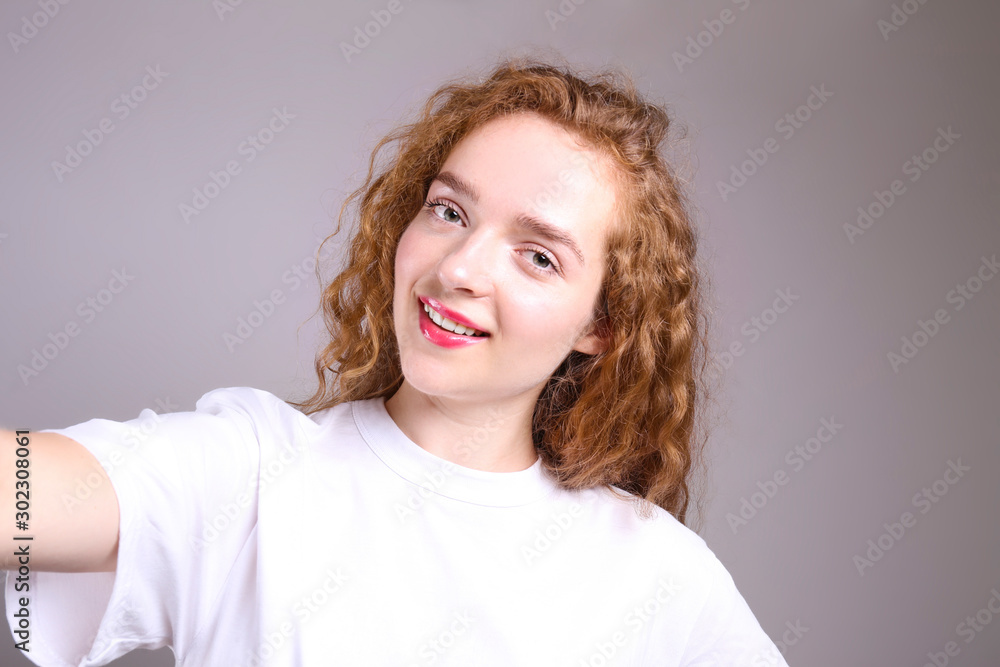 Young beautiful woman with long curly hair posing over isolated grey background. Portrait of teenage female model wearing white blank t-shirt, showing emotions. Close up, copy space. Young beautiful woman with long curly hair posing over isolated grey background. Portrait of teenage female model wearing white blank t-shirt, showing emotions. Close up, copy space.