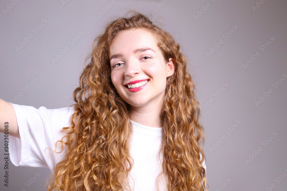 Young beautiful woman with long curly hair posing over isolated grey background. Portrait of teenage female model wearing white blank t-shirt, showing emotions. Close up, copy space. Young beautiful woman with long curly hair posing over isolated grey background. Portrait of teenage female model wearing white blank t-shirt, showing emotions. Close up, copy space.