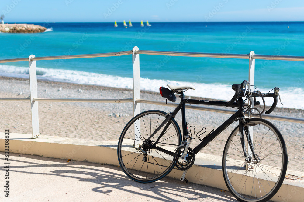 A bicycle sits parked along a ramp to the beach as a group of matching sailboats are blurred in the distance on the French Riviera in Menton, France