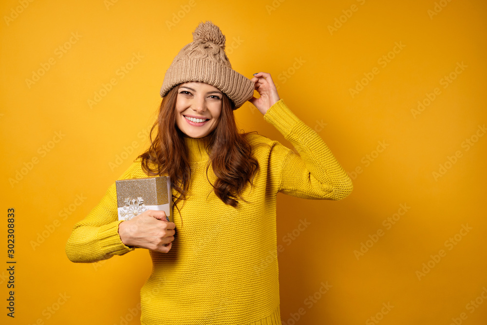 A beautiful brunette in a yellow sweater and hat stands on a yellow background with a snow-white smile and clutching a gift box. A beautiful brunette in a yellow sweater and hat stands on a yellow background with a snow-white smile and clutching a gift box.