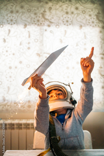 Fantasizing boy in astronaut helmet playing with paper plane at home photo