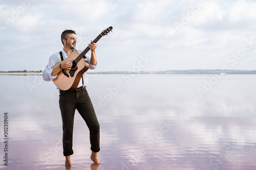 Passionate man in white shirt and suspenders playing guitar while standing barefoot in water by shore on cloudy day