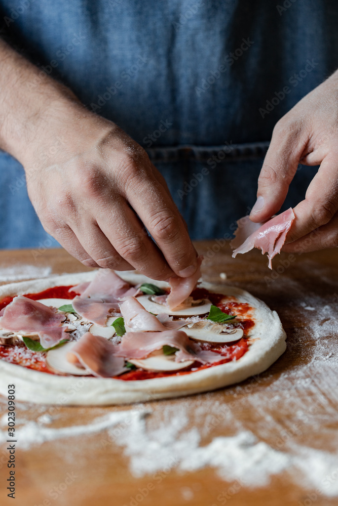 Unrecognizable chef in apron putting tasty ham on raw pizza while cooking on table in kitchen Unrecognizable chef in apron putting tasty ham on raw pizza while cooking on table in kitchen