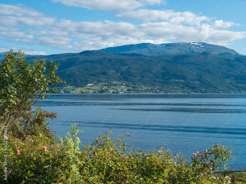 View over blue water of Sogndalsfjorden Fjord Sogndal with small village on the coast. Norway, nature and travel background. Early autumn blue sky background. View over blue water of Sogndalsfjorden Fjord Sogndal with small village on the coast. Norway, nature and travel background. Early autumn blue sky background.