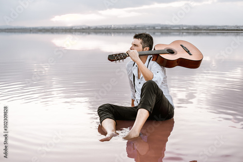 Wistful man in white shirt and suspenders carrying acoustic guitar and sitting on beach with pink water looking away surrounded with smooth sea reflecting majestic cloudscape