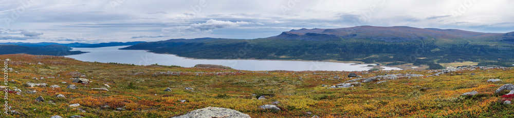 Panoramic landscape of wild nature in Sarek national park in Sweden Lapland with snow capped mountain peaks, river and lake, birch and spruce tree forest. Early autumn colors, blue sky white clouds.