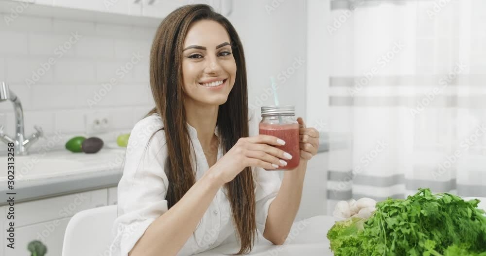 Attractive happy brunette woman drinking fresh smoothie in kitchen and smiling to camera, organic food dieting, keeping fit Attractive happy brunette woman drinking fresh smoothie in kitchen and smiling to camera, organic food dieting, keeping fit