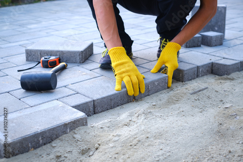 The master in yellow gloves lays paving stones in layers. Garden brick pathway paving by professional paver worker. Laying gray concrete paving slabs in house courtyard on sand foundation base.
