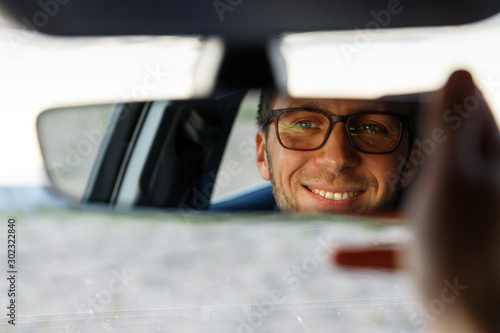 Cheerful joyfull man wearing glasses and adjusting mirror while sitting in his car  looking in reflection. soft focus. Emotions from driving a new car
