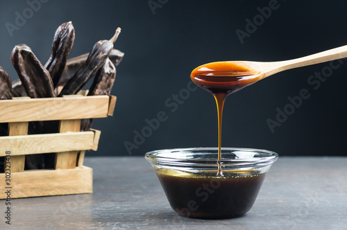 carob molasses in glass bowl and in wooden spoon and carob pods on rustic background, locust bean healthy food, Ceratonia siliqua ( harnup ) photo
