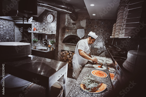 Italian chef in uniform is adding tomato sauce for pizza at the kitchen.