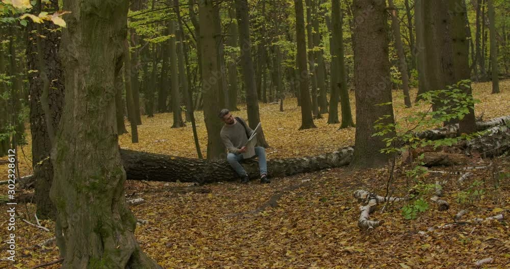 Handsome smiling Caucasian man sitting on the trunk in the autumn forest and looking at map. Male tourist searching the best way to go. Cinema 4k footage ProRes HQ. Handsome smiling Caucasian man sitting on the trunk in the autumn forest and looking at map. Male tourist searching the best way to go. Cinema 4k footage ProRes HQ.