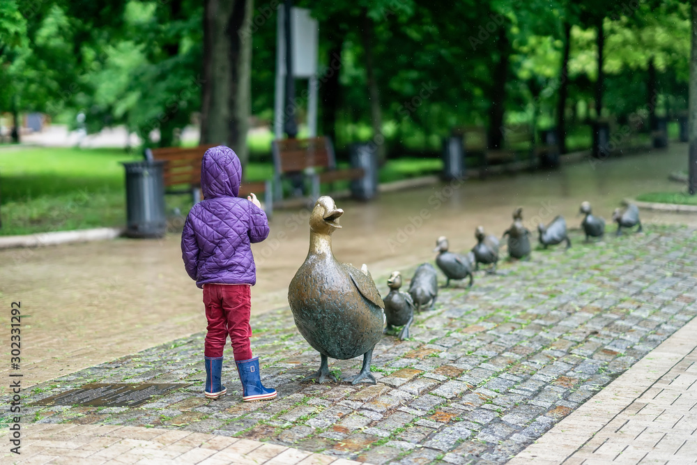 Fototapeta premium Unrecognizable little girl with her back to us counts the number of little ducklings of mothers duck, sculpture in the park