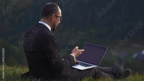 Successful Businessman Makes A Deal On The Phone Holding Laptop On His Lap While Sitting On A Hill
