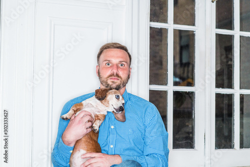 Slika na platnu Young American Man with beard, wearing blue patterned shirt, sitting at home by window in New York City, holding Dachshund dog, relaxing