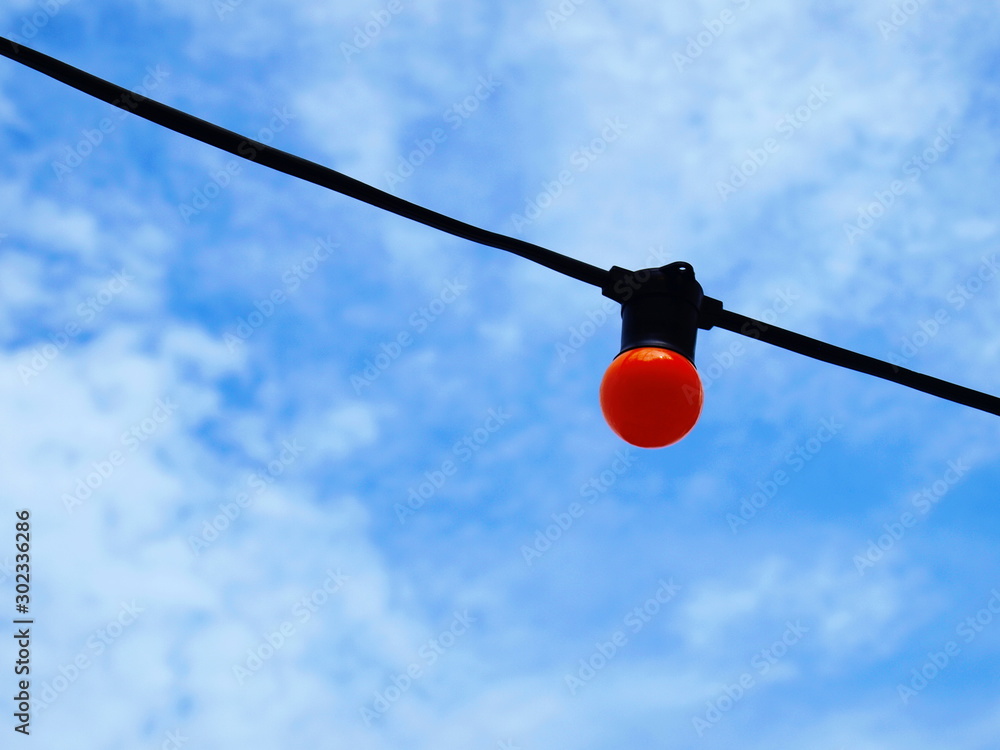 A round orange bulb close up hanging on a black wire On the blue sky background with white clouds With copy space. A round orange bulb close up hanging on a black wire On the blue sky background with white clouds With copy space.