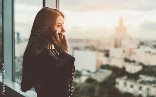 Sideview of an adult beautiful caucasian businesswoman talking on the phone while standing next to a big window of her business office with a copy space area on the right and the cityscape outside photo