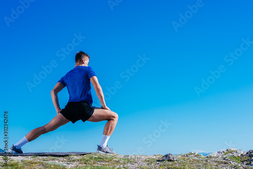 Blonde athlete wearing blue dry-fit shirt stretching and preparing for a workout while standing on top of a cliff which has an amazing view.