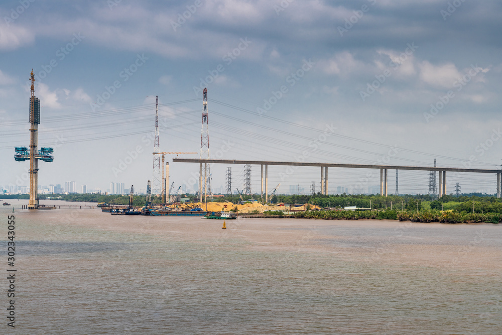 Long Tau River, Vietnam - March 12, 2019: Phuoc Khanh Bridge under construction, not yet over river, but on ramp. Tall stong pylon raises out of brown water. Under blue cloudscape, yellow sand and gre