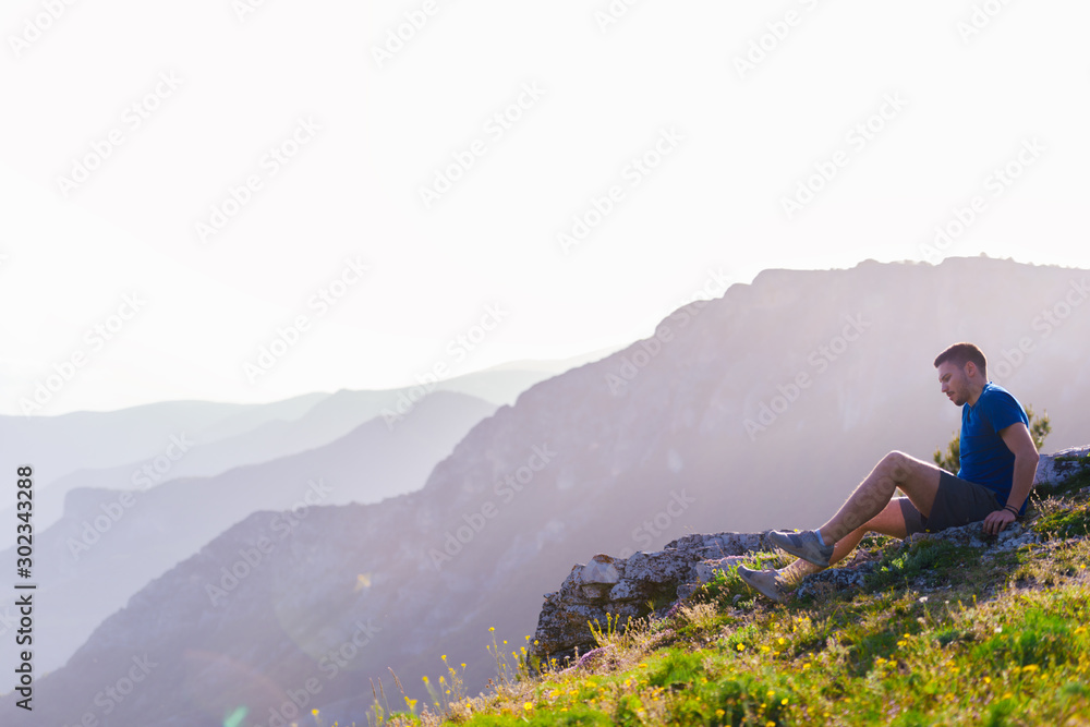 Adventurous man sitting on top of a mountain and enjoying the beautiful view, while looking downhill at the blue river and amazing mountain line..