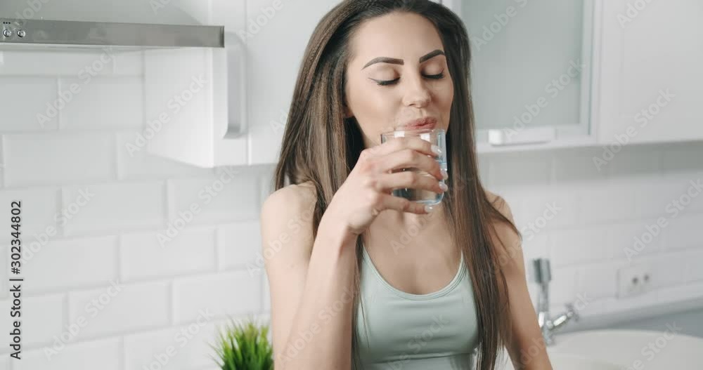 Sporty brunette woman drinking water after workout standing in kitchen, maintaining water balance Sporty brunette woman drinking water after workout standing in kitchen, maintaining water balance