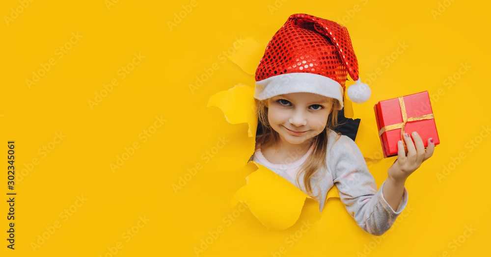 A cute little smiling girl in a red Santa hat looks out of a hole in yellow paper and holds a box with a gift in her hand. Christmas and New Year concept. Bright coloured background, copy space. A cute little smiling girl in a red Santa hat looks out of a hole in yellow paper and holds a box with a gift in her hand. Christmas and New Year concept. Bright coloured background, copy space.