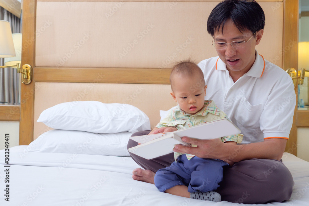 Smiling father and Cute little Asian 18 months / 1 year old toddler baby boy reading bedtime story book, sitting on bed at home, Dad and son spending time together, Parents reading to child concept Smiling father and Cute little Asian 18 months / 1 year old toddler baby boy reading bedtime story book, sitting on bed at home, Dad and son spending time together, Parents reading to child concept