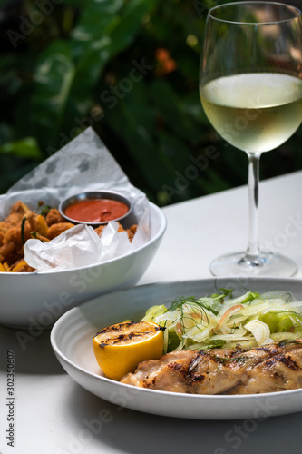Grilled white fish with lemon and greens on a side and fried calamari rings with glass of white wine outside restaurant interior