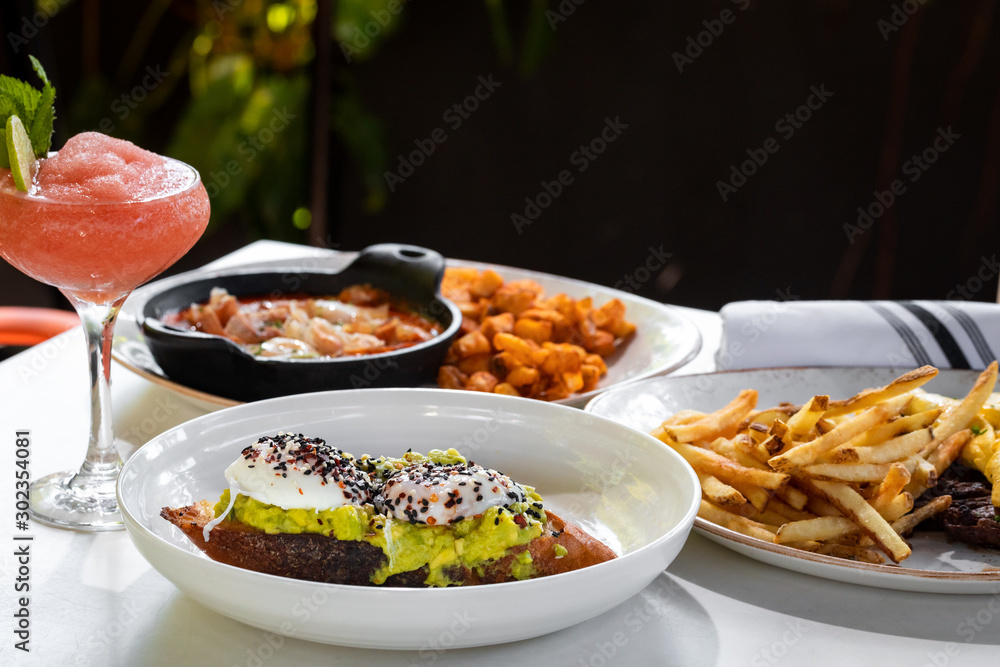 Business lunch of  jamon and ham skillet, toast with avocado and egg, grilled beef with french fries and pan with brussels sprouts with drinks outside a restaurant in a sunny day 