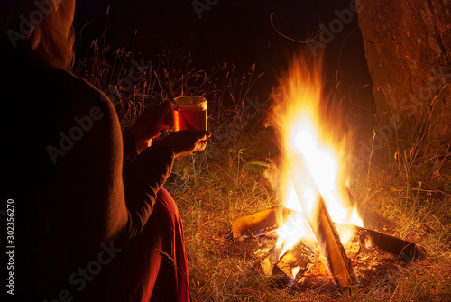 A young girl is drinking tea by the fire in the night forest.
