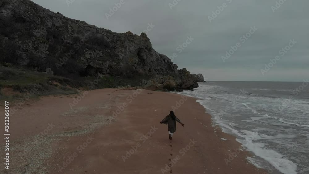 A girl in a coat with a scarf flying in the wind walks along the sandy beach in the evening A girl in a coat with a scarf flying in the wind walks along the sandy beach in the evening