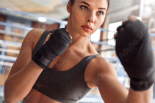 Boxing. Woman boxer with bandage around hands exercise kick near ring serious close-up