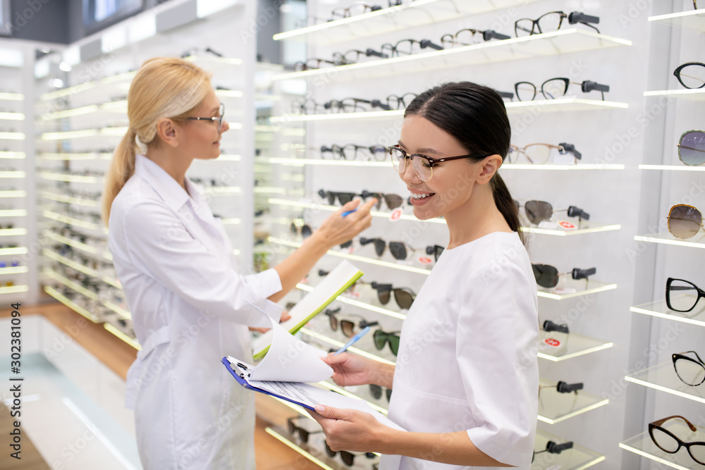 Dark-haired eye doctor smiling while making notes after ordering glasses Dark-haired eye doctor smiling while making notes after ordering glasses