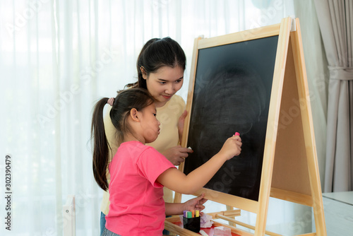 Asian mother playing with her daughter drawing together with color chalk in blackboard at table in living room at home. Parenthood or love and bonding expression concept.