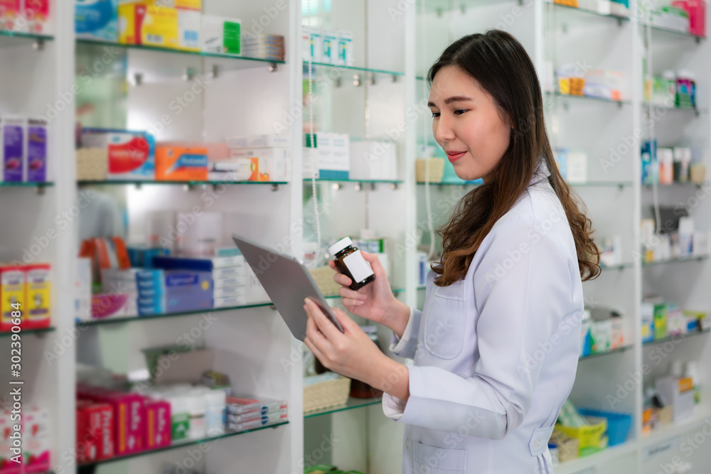 Confident Asian young female pharmacist with smile holding a medicine bottle and searching that product in digital tablet database in the pharmacy drugstore. Medicine, pharmaceutics, health care
