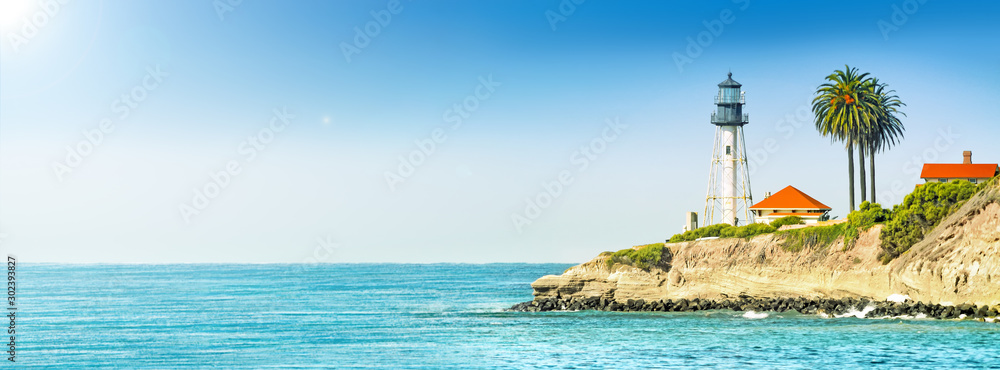 lighthouse tower on cliff in ocean landscape against stormy sea water and blue sky background. Panorama ultra wide side view of marine light beacon with palm tree located on rock. Copy space banner - obrazy, fototapety, plakaty 