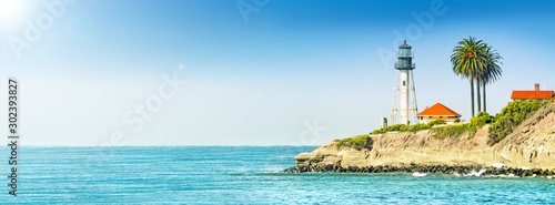 lighthouse tower on cliff in ocean landscape against stormy sea water and blue sky background. Panorama ultra wide side view of marine light beacon with palm tree located on rock. Copy space banner