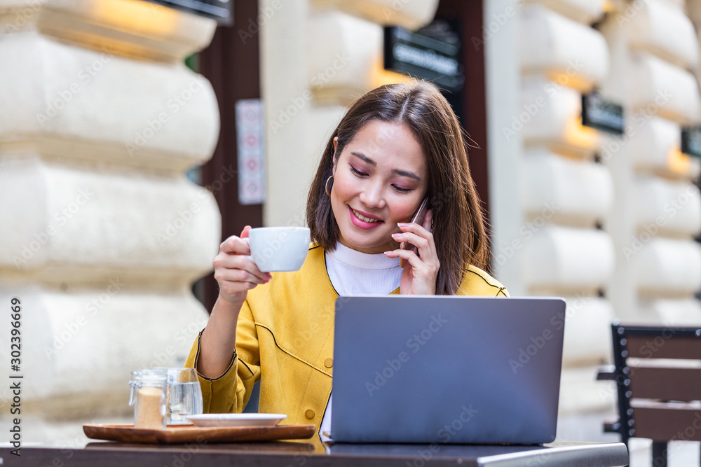 Young Asian woman sitting in coffee shop at wooden table, drinking coffee and using smartphone.On table is laptop. Girl browsing internet, chatting, blogging.