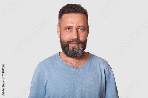 Studio shot of middle aged man with beard dresses grey casual t shirt, posing isolated over light background, attractive male looking directly at camera with calm facial expression. People concept. © sementsova321