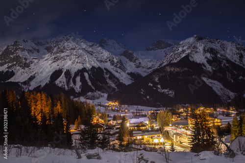 Snowy winter mountains at night with town lights in a saddle and forests. 